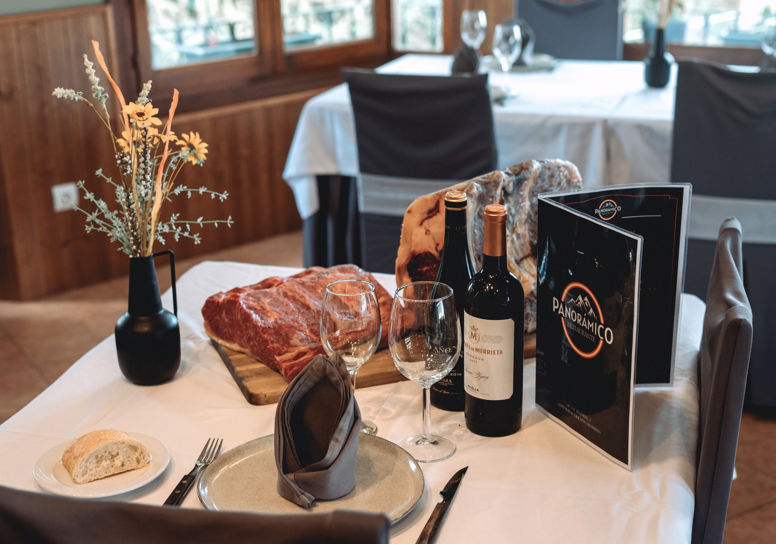 Vista de una mesa engalanada con vino tinto, copas, carne fresca, la carta y un jarrón con flores secas.